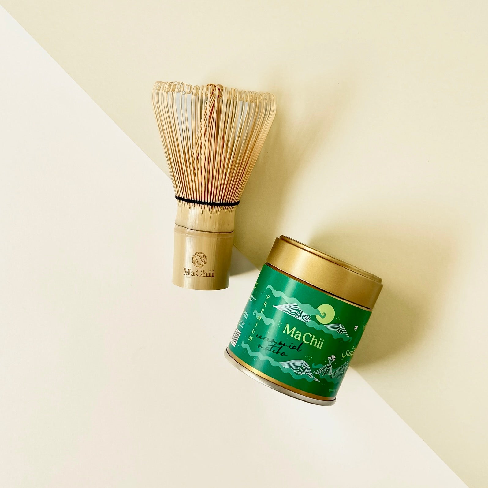 organic ceremonial matcha premium tin placed next to a bamboo matcha whisk with 120 prongs. the logo of machii tea is on the tin in green as well as the whisk