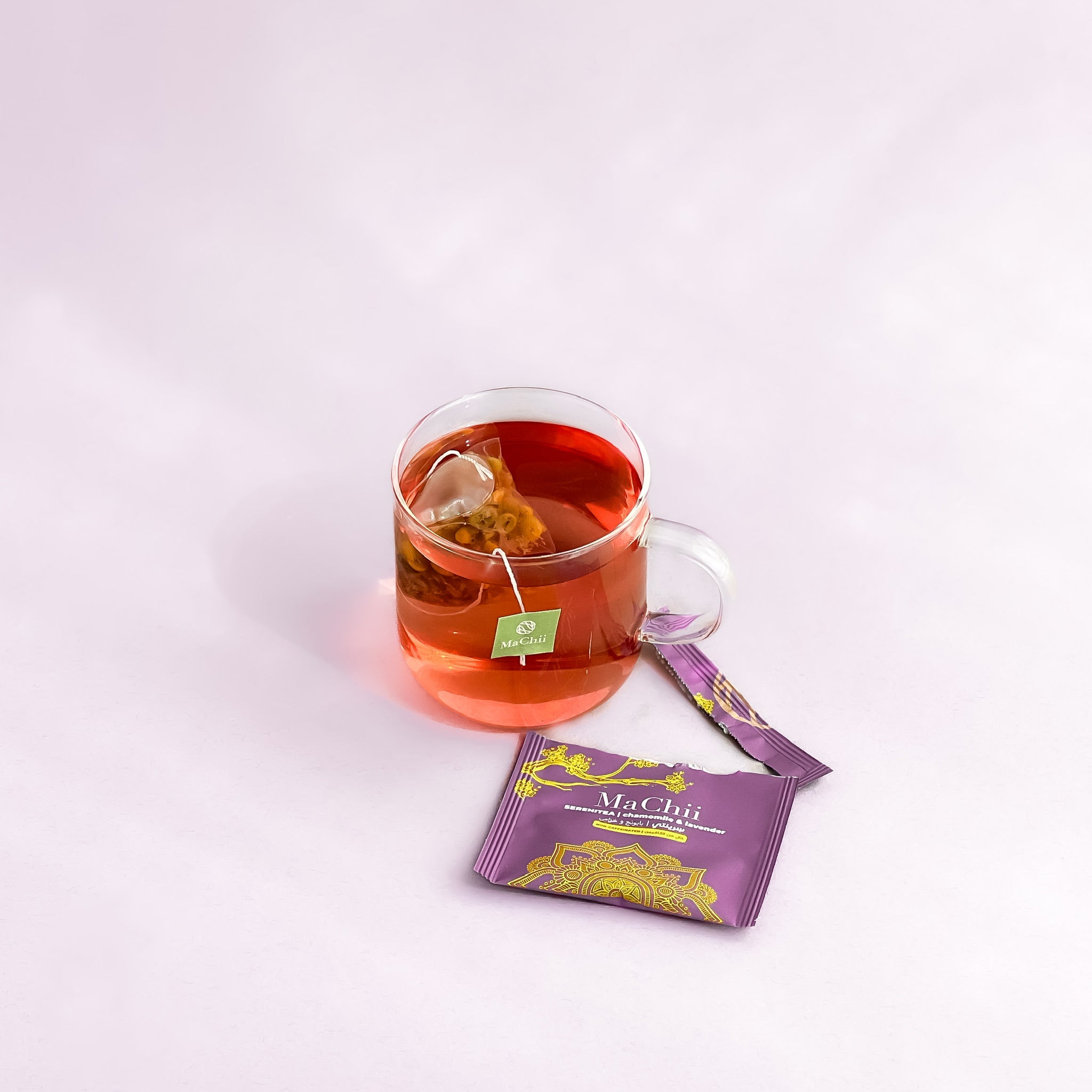 SereniTea infusing in a glass mug with the tea tag hanging off the side of the cup. The envelope is teared open. the infusion is with an organic silk tea of chamomile, hibiscus and lavender. 