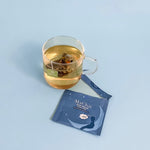 sweet dreams herbal blend chamomile in tea silk biodegradable tea bag on top of an envelope package with a picture of a sleeping man on a moon at night. the tea bag is brewing in a cup and is yellow