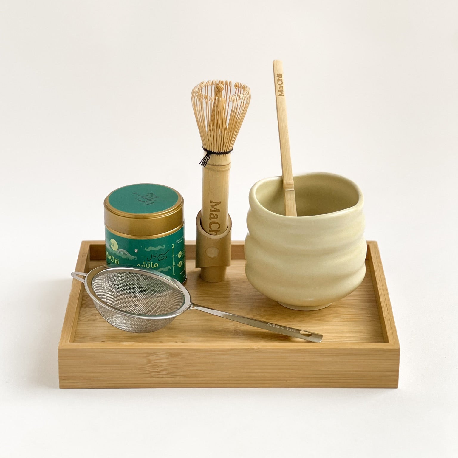 Tea-making set with a ceramic cup, whisk, scoop, and tea canister on a wooden tray.