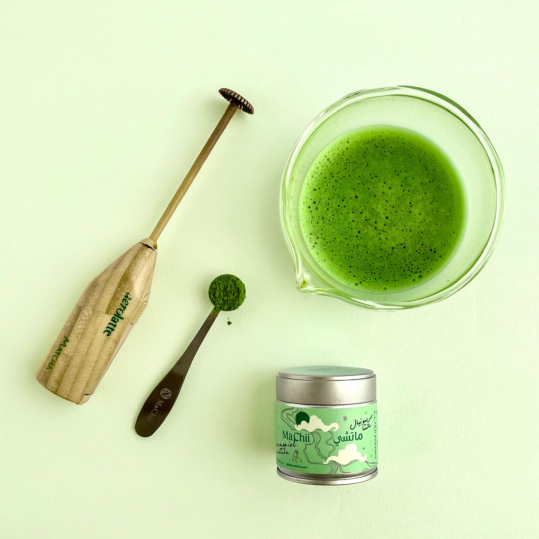 modern matcha latte bundle with an aerolatte electric matcha whisk, a silver matcha tin, a matcha tea spoon and a glass bowl with spout filled with green powder.