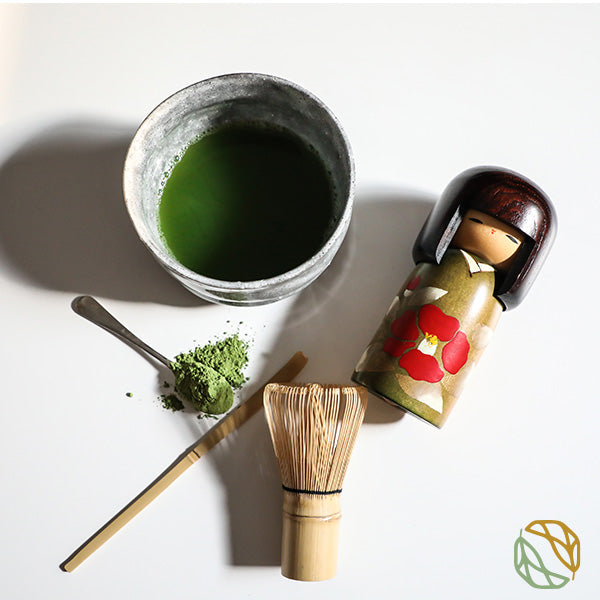 organic ceremonial grade premium matcha from machii tea. it is made in a matcha bowl with a Japanese doll on the side. bamboo match scoop with powder in the background. 